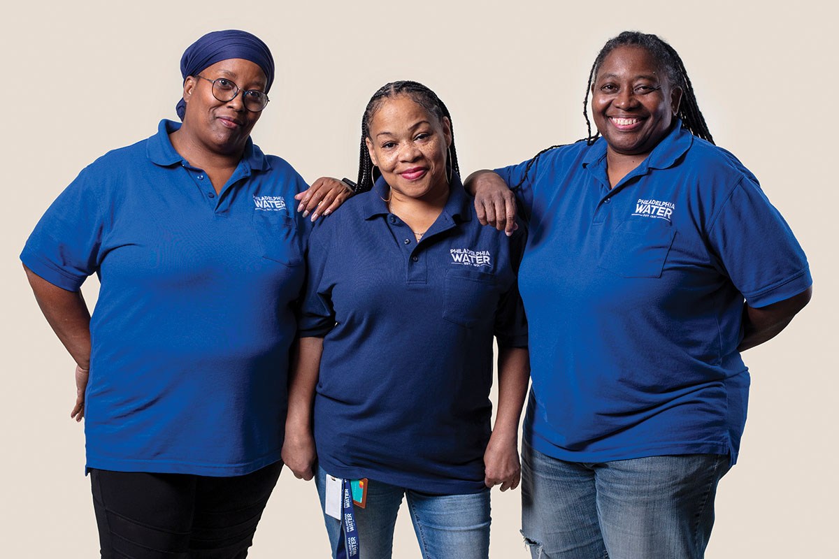 Three Contact Center reps in blue PWD polo shirts and jeans stand and pose, the two taller women on either side resting a shoulder on the one in the center. One wears a dark blue wrap over her hair and wire framed glasses, the other two have long dark hair in small braids.