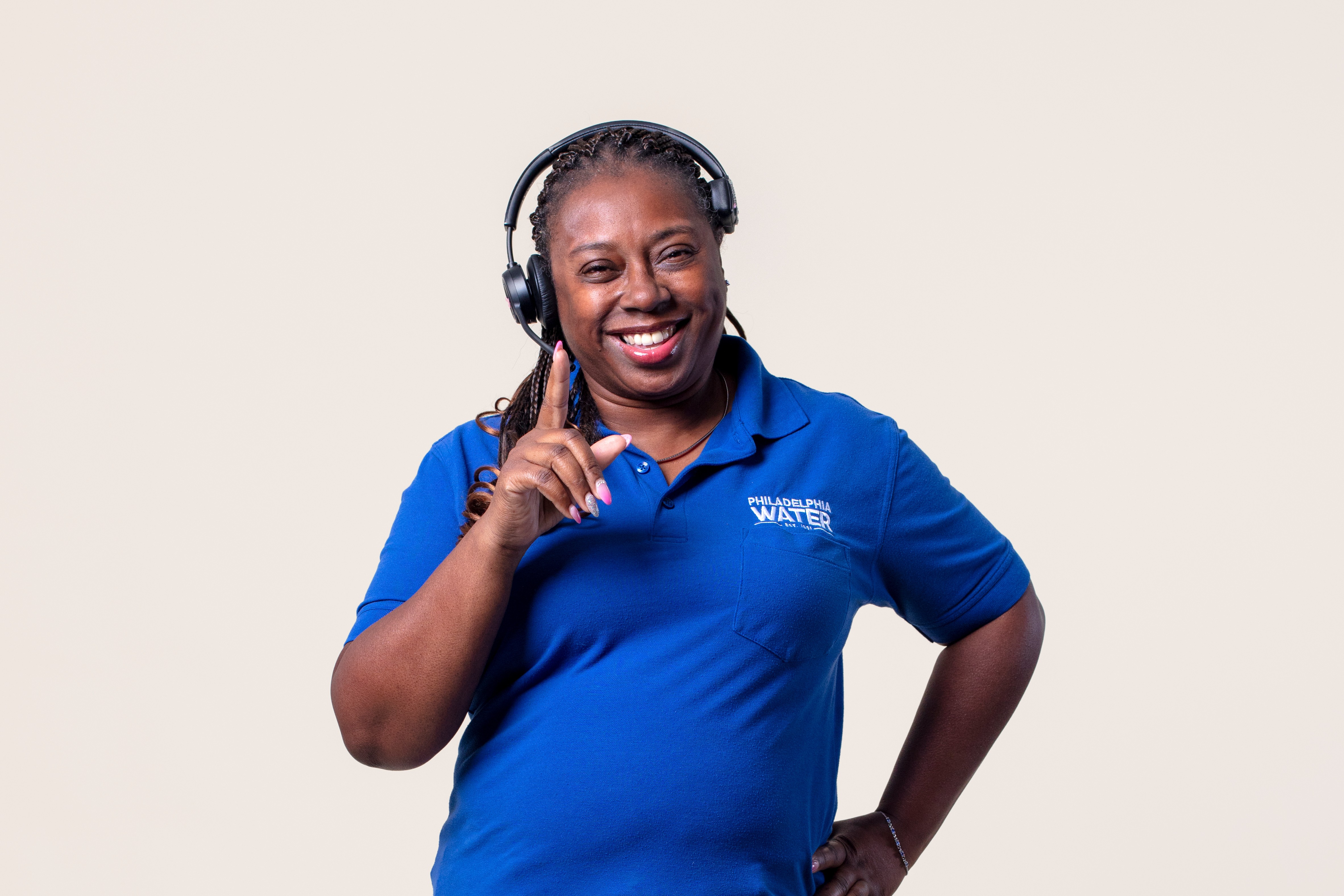 A smiling contact center representative with dark skin and hair in long braids, wearing a bright blue PWD polo shirt and black headset, holds up one finger, the other hand on her hip.