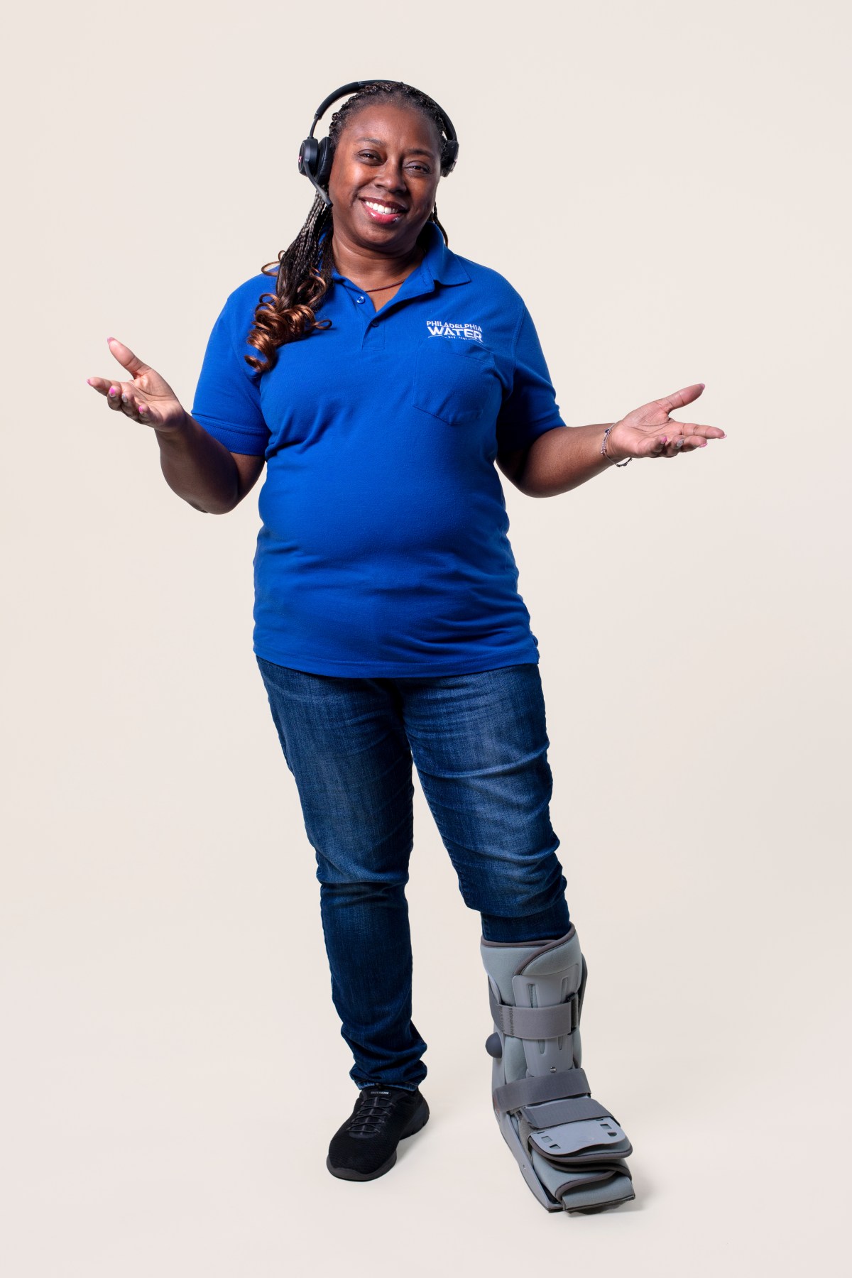 A contact center rep wearing a headset, blue PWD polo, jeans, and her foot in a boot, stands with arms extended in welcome.