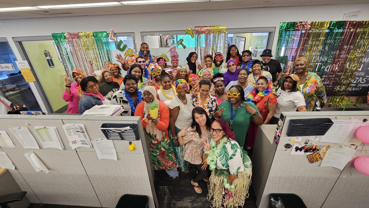 Representatives from the PWD Contact Center stand together in the office wearing festive, colorful clothing. Behind them is a wall draped with curtains of colorful metallic material and a banner that reads Luau Party