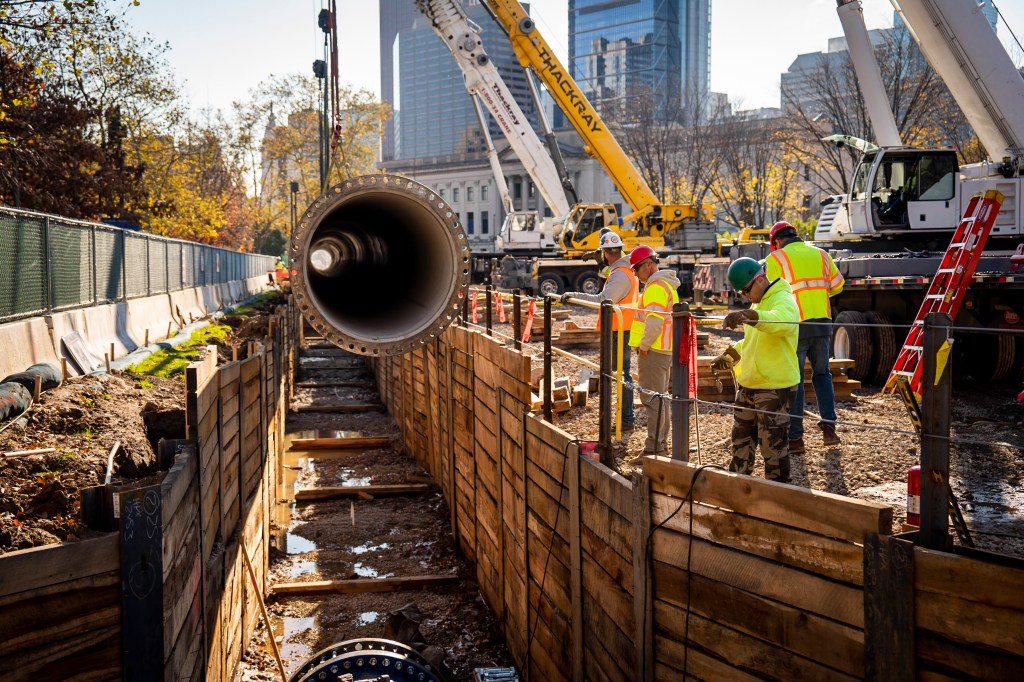 A section of new water main is lowered into a trench stabilized with wood plank walls holding up the sides. We’re looking down the length of the trench and into one end of the pipe, which has a raised ring on the end, with holes for bolting it onto the next section all the way around it. Two crane arms are visible in the top right corner, and behind them we see a bit of the Franklin Institute, One and Three Logan Square, and other buildings in the background. Trees with fall foliage line the left side, behind the construction fencing.