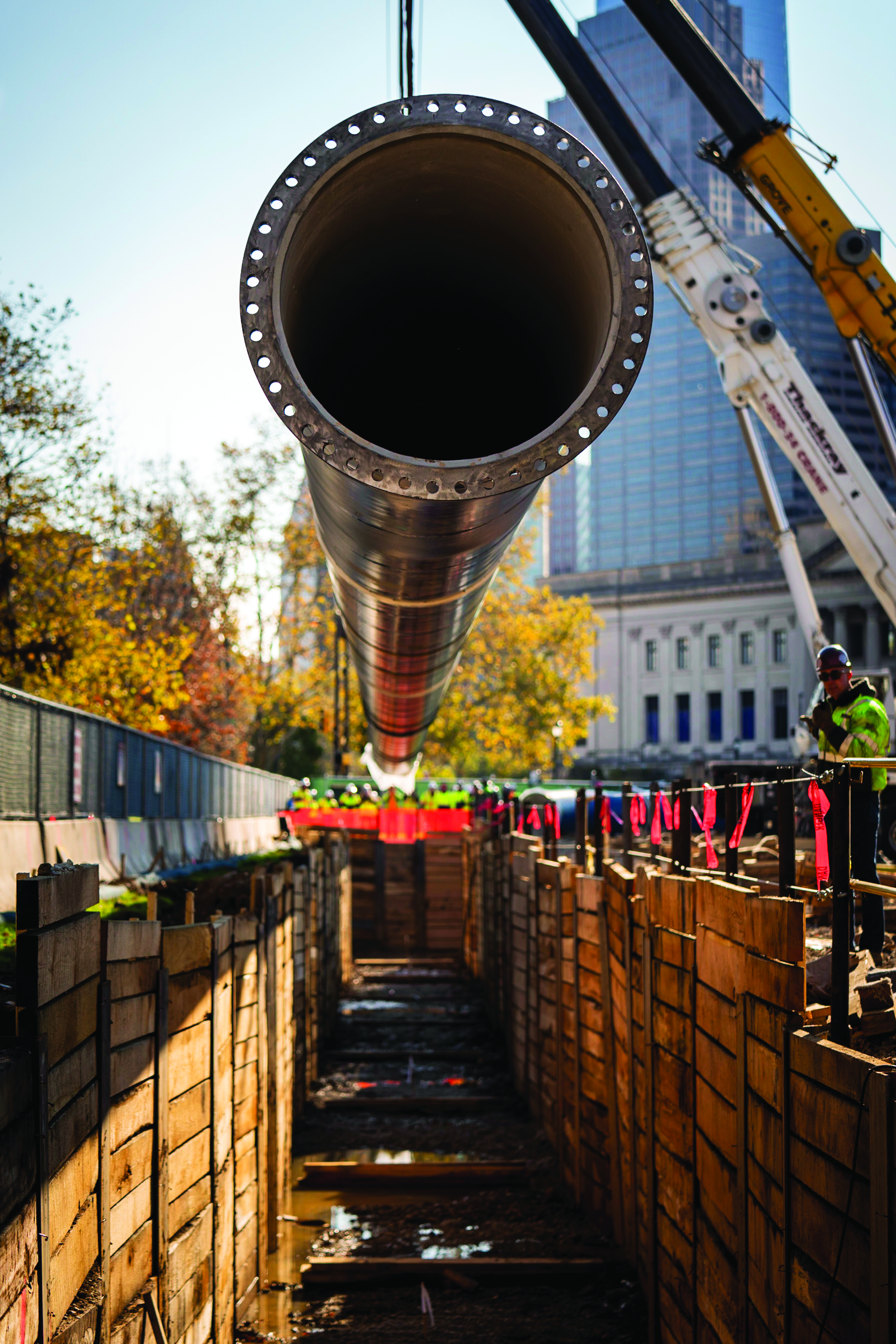 A section of new water main is lowered into a trench stabilized with wood plank walls holding up the sides. We're looking down the length of the trench and into one end of the pipe, which has a raised ring on the end, with holes for bolting it onto the next section all the way around it. Two crane arms are visible in the top right corner, and behind them we see a bit of the Franklin Institute, One and Three Logan Square, and other buildings in the background. Trees with fall foliage line the left side, behind the construction fencing.