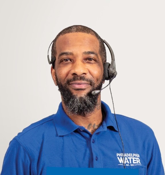 a PWD contact center representative with close cropped black hair, beard, and mustache, wearing a phone headset and blue polo shirt with the Philadelphia Water Department logo is visible from the chest up looking straight at the camera with a subtle smile.