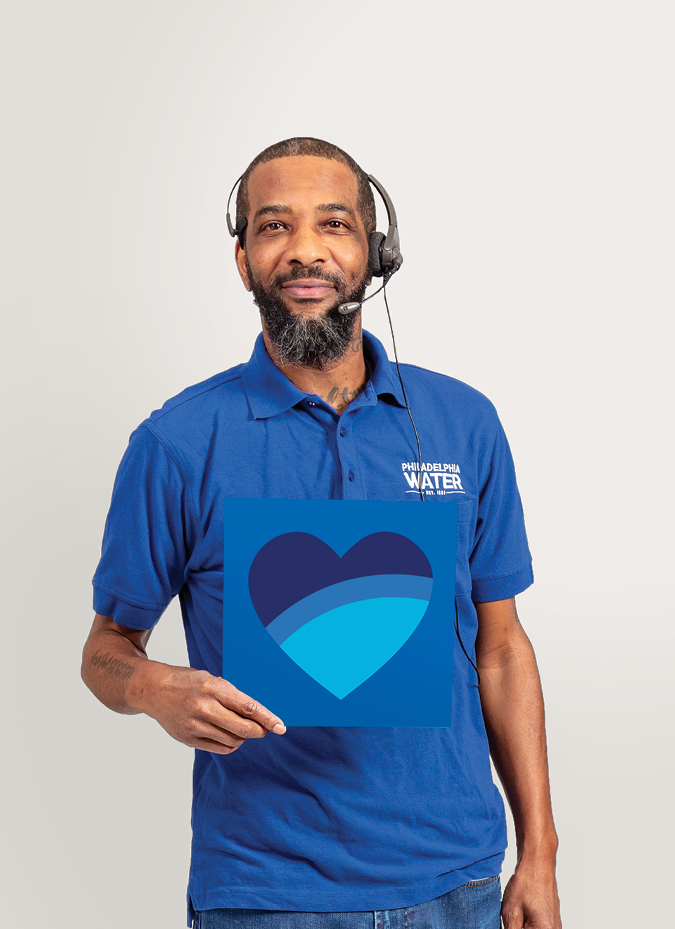 A PWD Customer Service Representative holds up a card with a heart on it, shaded with 3 shades of blue like the PWD logo. He is wearing a blue PWD polo and a headset, and has warm brown skin and dark, close cropped hair and a short beard.