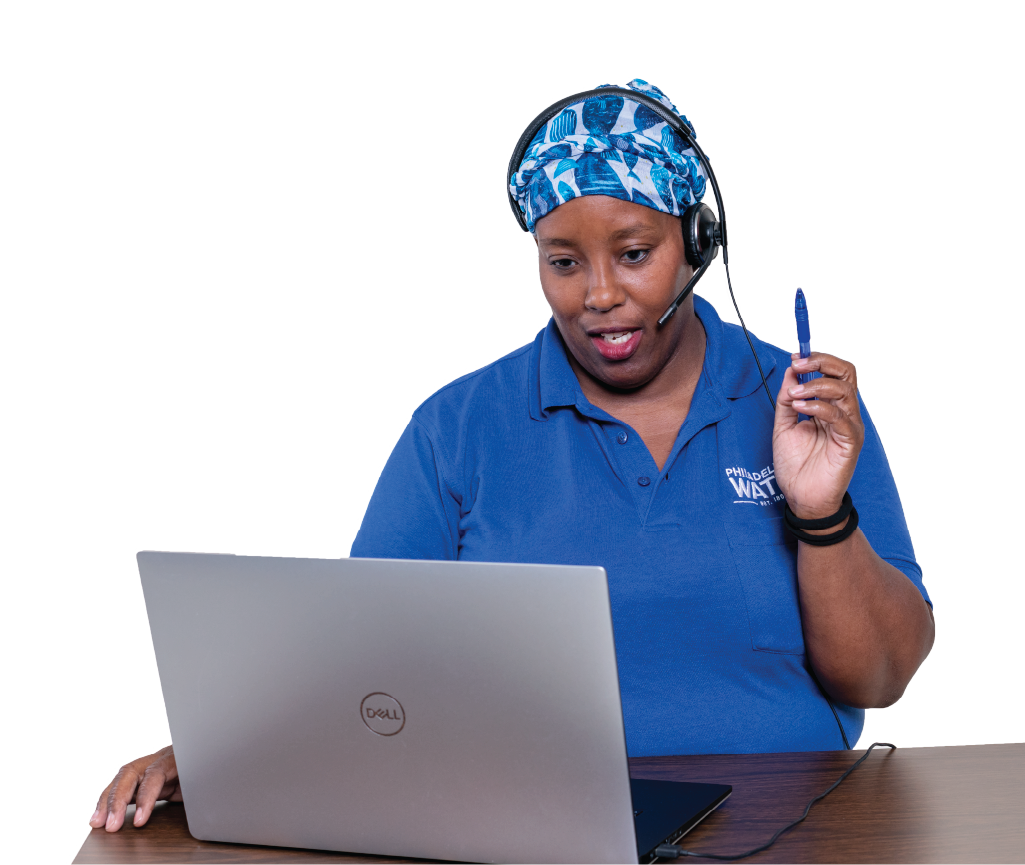 A contact center representative wearing a blue PWD polo, a headwrap with a vibrant blue and white fish pattern, two black bracelets, and a phone headset sits at a desk, looking at a laptop, and seems to be speaking with a customer, mouth partially open and a pen held in the air as if in thought.