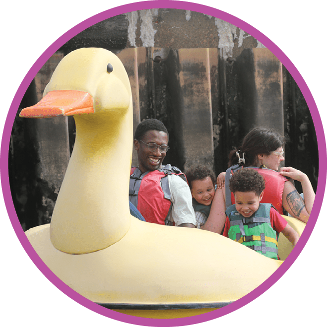 A family wearing life jackets laugh as the small children attempt to help operate a paddle boat shaped like a giant duck.