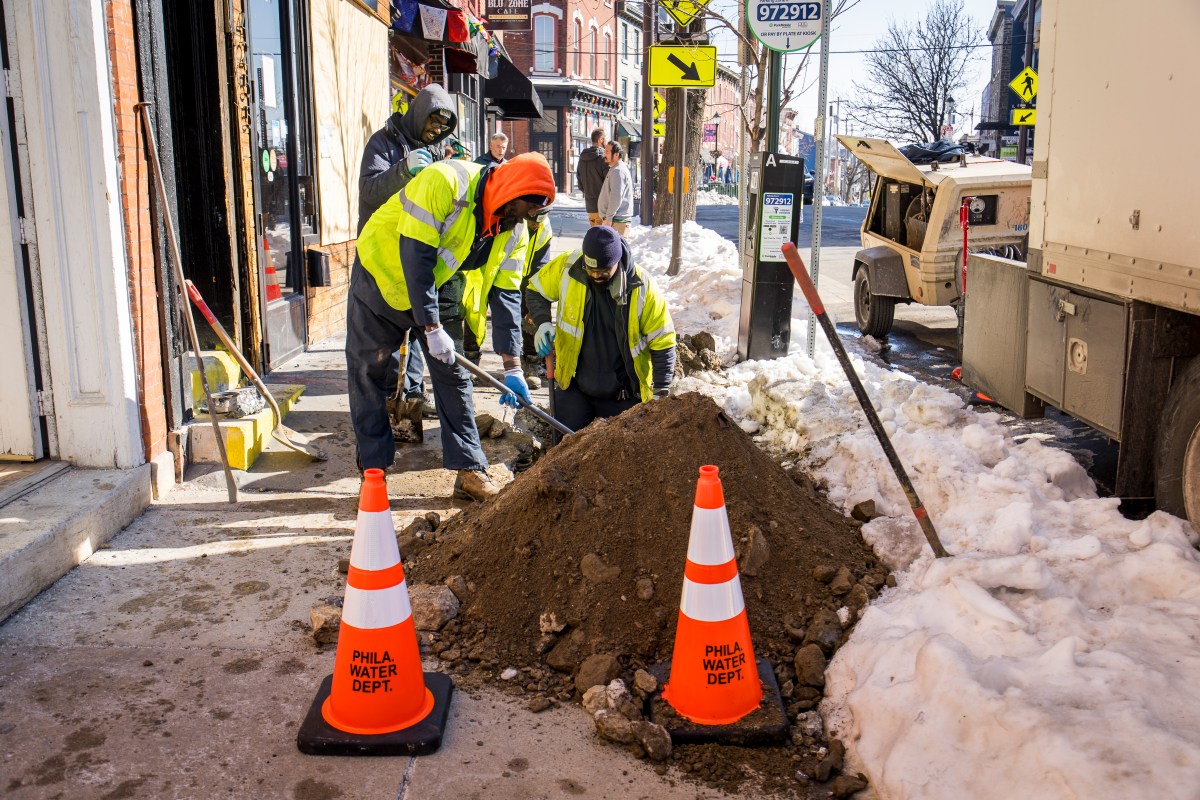 A crew wearing reflective safety vests over winter gear dig a hole in the middle of a sidewalk, one knee-high in it with the others standing on the edges. There are storefronts visible on the left, a long mound of shoveled snow on the right, and two orange cones and the mound of excavated dirt in the foreground.