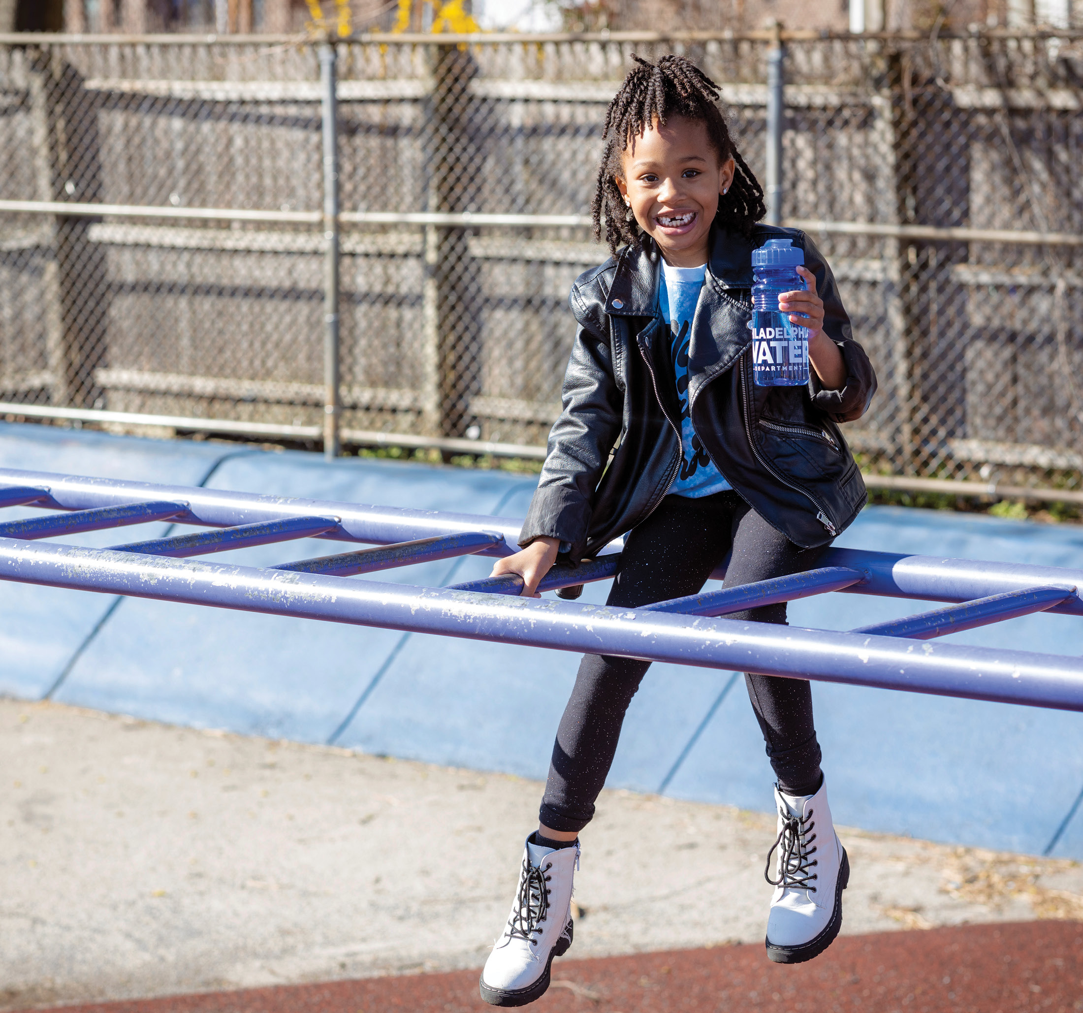 A young girl with a huge smile showing off a missing tooth as she sits on top of the monkey bars at Ellwood Elementary School, holding up her reusable PWD water bottle. She has brown skin, dark hair in shoulder length braids, half pulled up on top of her head, and is wearing a black leather jacket over a blue tie-dye tshirt with black leggings and white lace-up boots.