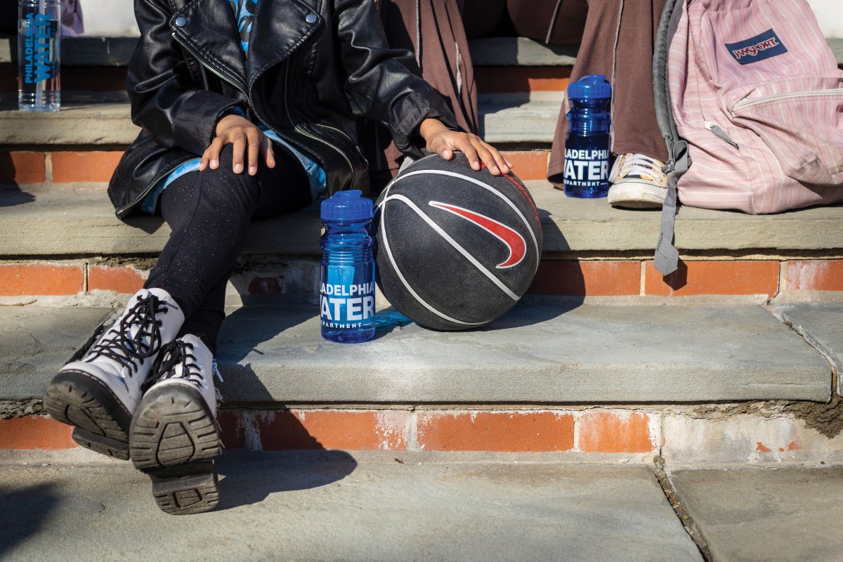 Two kids are sitting on steps with thick slate treads and brick risers. One is visible from the chest down, wearing a black leather jacket over a blue t-shirt and sparkly black leggings, with white lace-up boots, with her legs crossed, one hand on her knee and the other on a black basketball. Another, sitting on a higher step, can only be seen from below the knee, wearing brown pants and yellow Chuck Taylors, with a pink backpack leaning against a leg. Both have blue translucent water bottles with the PWD logo on them, mostly full of water. The shadow of a third person and a clear PWD water bottle can be seen in the corner.