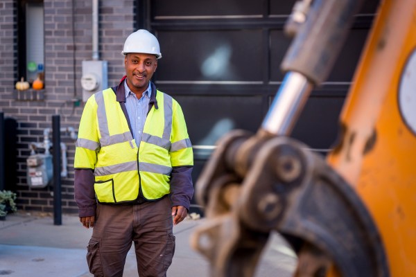 PWD employee wearing a hard hat and safety vest over a blue striped button-down shirt and cargo pants smiles at a lead service line replacement site. The bright yellow arm of an excavator is out of focus in the right foreground.