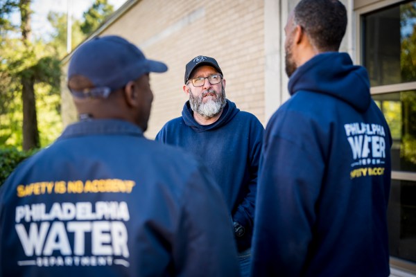 3 PWD employee having a conversation, all wearing dark blue PWD hoodies or jackets, two wearing baseball caps. One in the center faces the camera while the other two are facing him, and we can see "Safety is no accident" and the PWD logo on their backs.