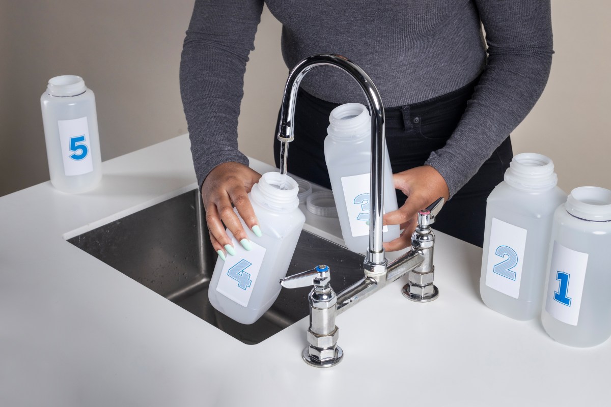 Hands with warm brown skin and long mint-colored fingernails filling sample collection bottles from a sink faucet.