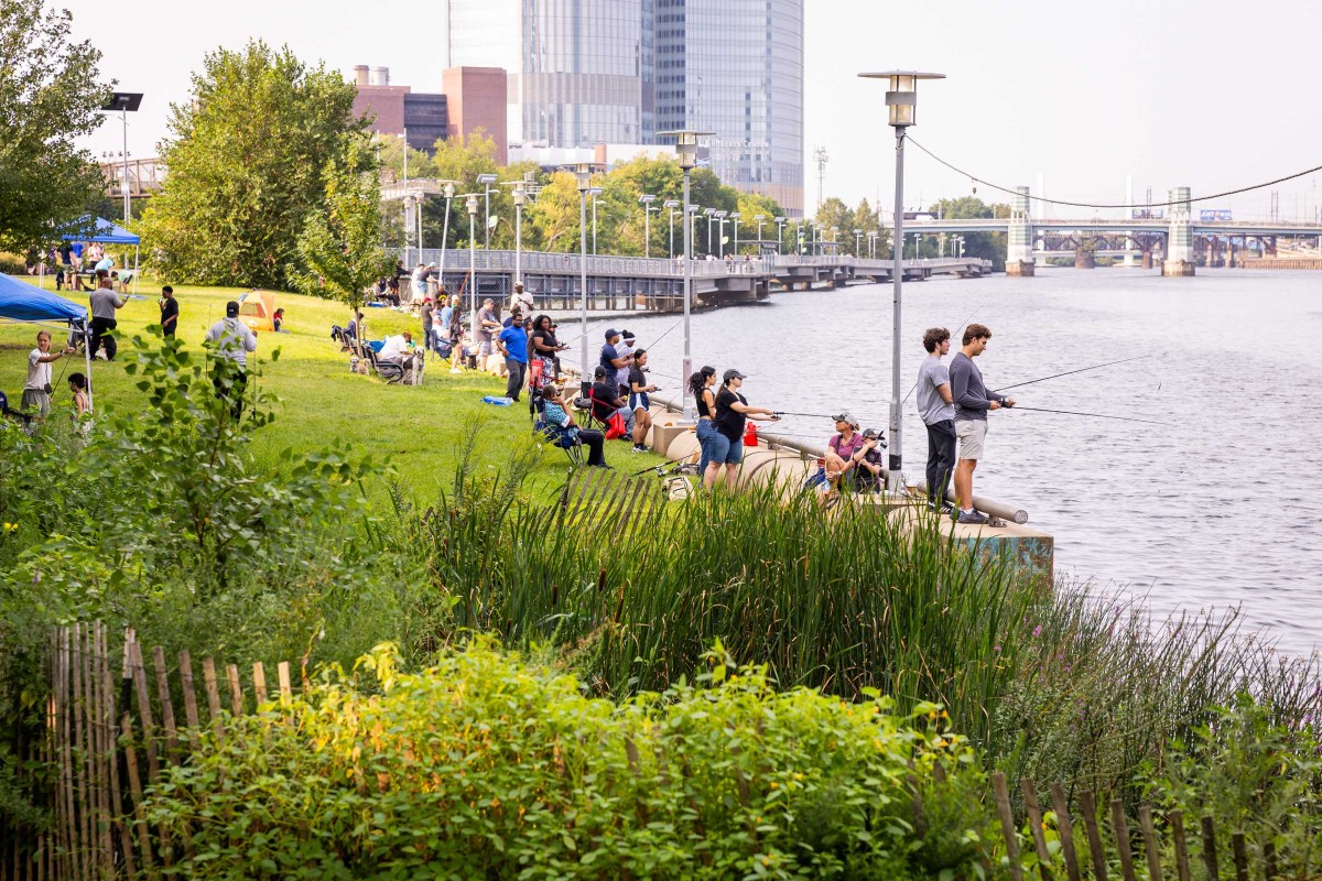 Photo of Fishing Fest 2025, where anglers of all ages sit or stand along the banks of the Schuylkill, many with rods extended out over the river.