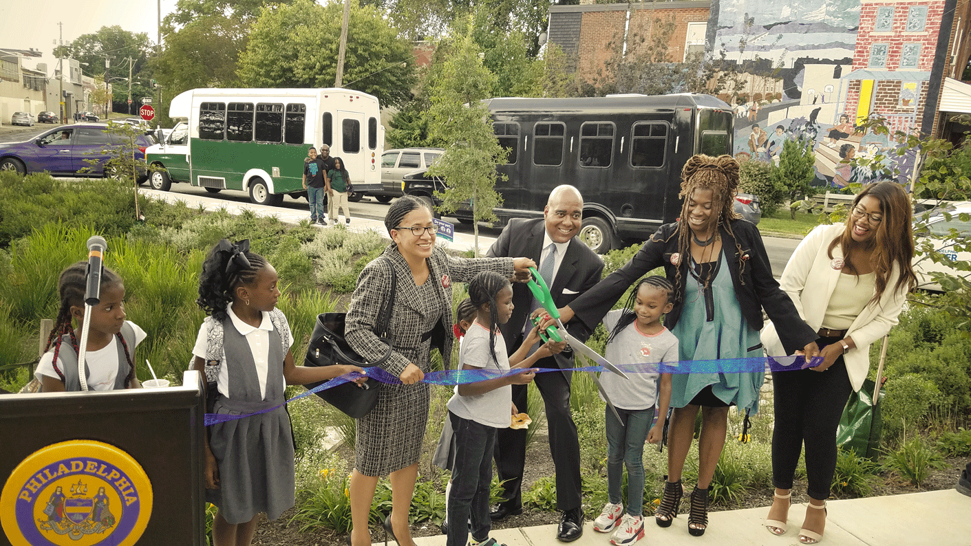 a row of PWD employees, community members, and children hold a ribbon stretched out in front of a newly completed rain garden, with several helping to hold very large scissors, poised to cut it