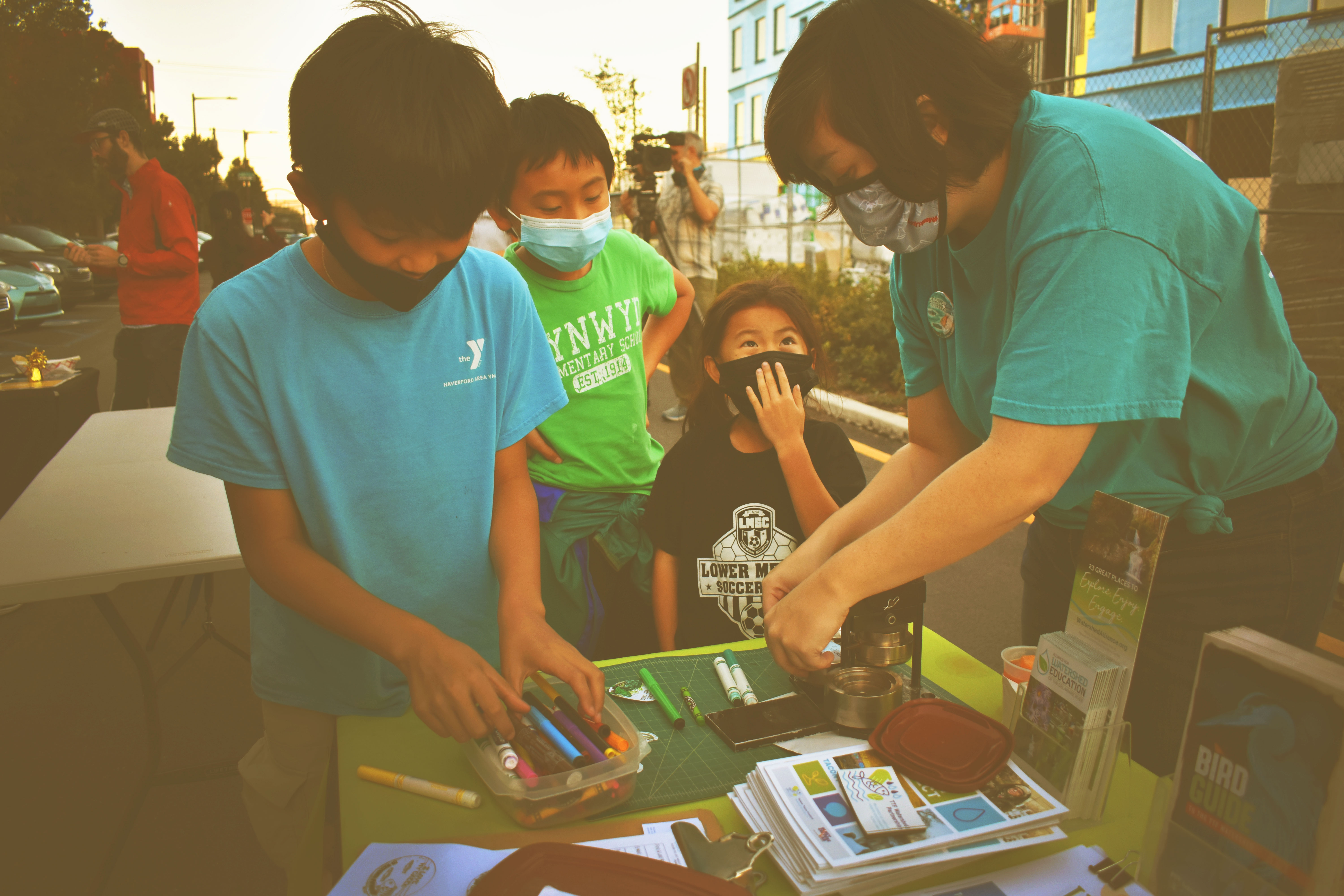 Three children in t-shirts and face masks gather around the Watershed Alliance table to color pins proclaiming their love for their creek at the Green City, Clean Waters 10 Year Celebration, as an adult operates the button-making machine. Other guests and news crews are visible in the background.