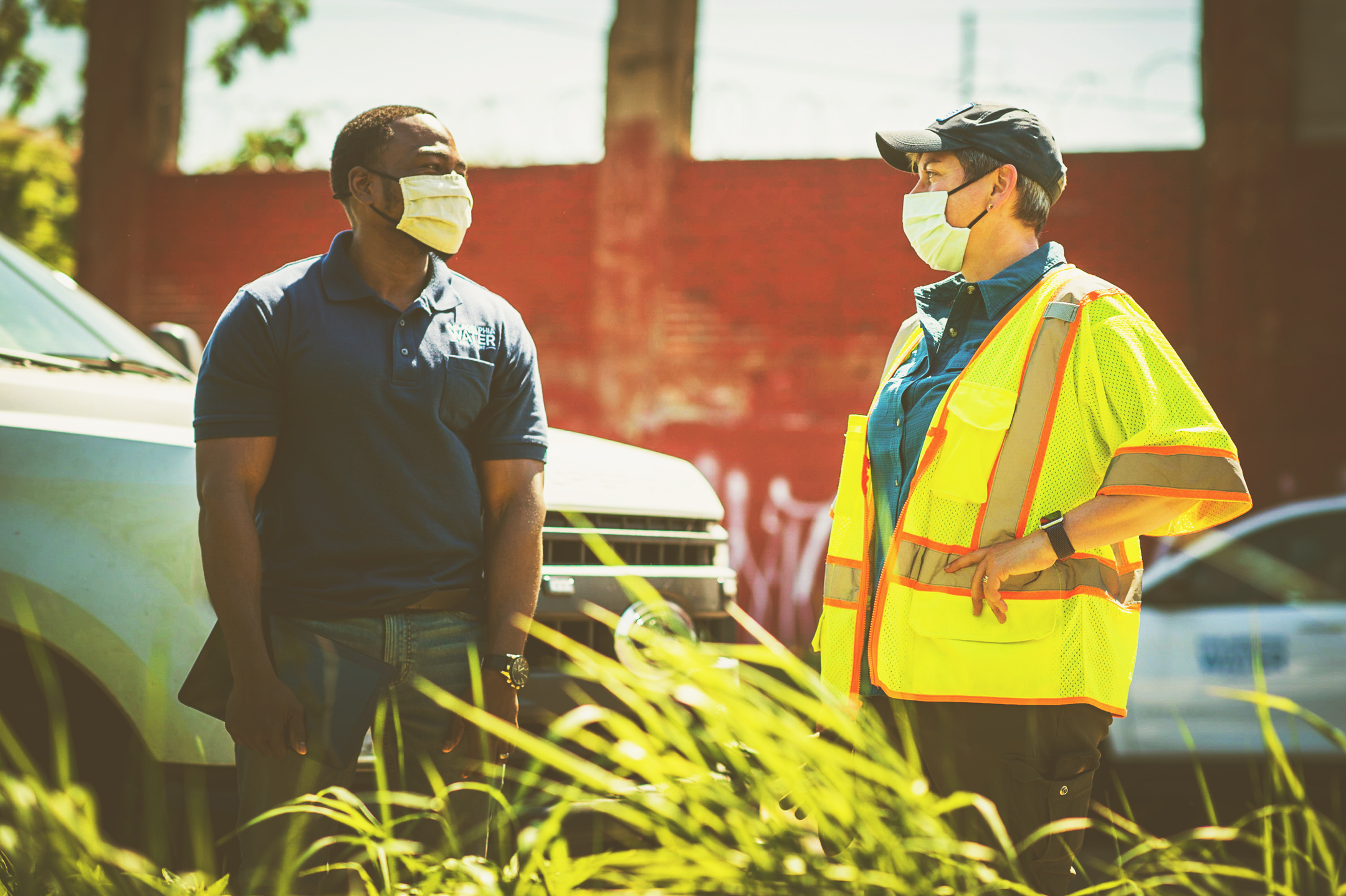 Two people in face masks stand outdoors, having a conversation. The tops of plants are visible in the foreground and two white vehicles are partially visible behind them, one showing the PWD logo on the side, with a brick wall in the background. One of the people has dark skin and close-cropped black hair, is wearing a navy blue PWD polo shirt and jeans, and holds a tablet with the screen darkened in one hand. The other has light skin and short salt and pepper hair, and is wearing a teal button-down shirt, dark cargo pants, a yellow safety vest, and a blue PWD baseball cap.