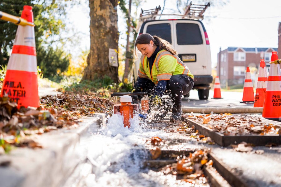 A young woman with a brown ponytail squats at a curb working with monitoring equipment. A white van is behind her and water froths in front of her in the gutter. Fallen orange leaves cover the curbside and the road in front of the worksite. On both sides of the worksite there are orange road cones with Phila Water Dept stenciled on them arranged for safety
