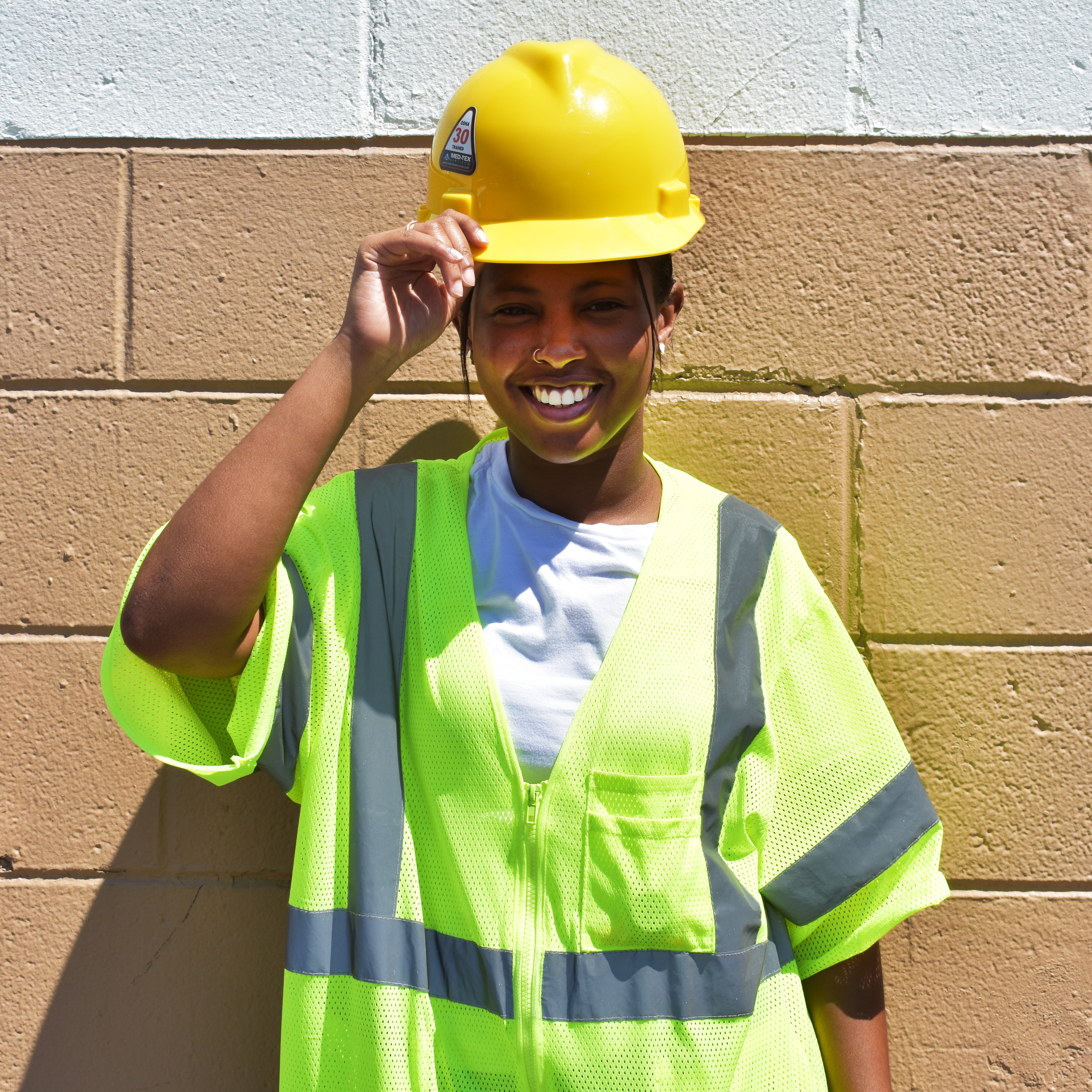 Jasmine started at PWD as an Apprentice in the Safety unit. She is pictured here with a big smile, holding the brim of a yellow hard hat worn over her pulled-back dark hair. She is wearing a neon yellow-green safety vest over a white t-shirt and standing in front of a concrete block wall mostly painted tan (the top row visible is painted white).