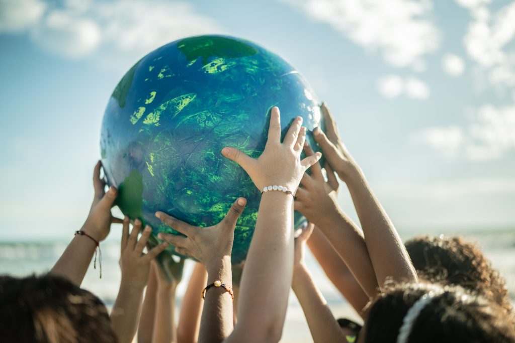 a bunch of children’s hands reaching up, holding up a ball, about basketball-size, that looks like a globe, with a light blue sky and whispy clouds visible in the background