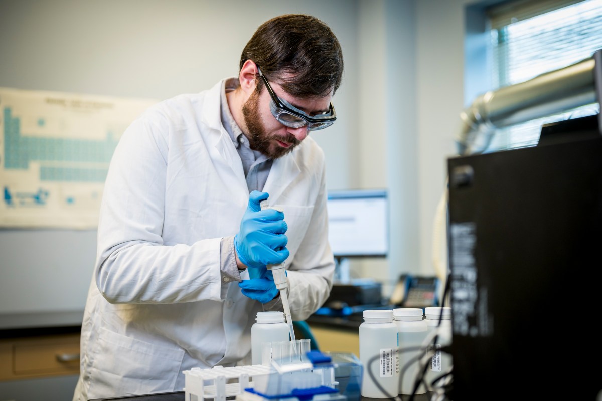 A PWD scientist with short brown hair and beard, wearing safety goggles, a white lab coat, and blue disposable gloves, leans over a workbench, carefully using a pipette to transfer water from a sample collection bottle to a row of test tubes for analysis.