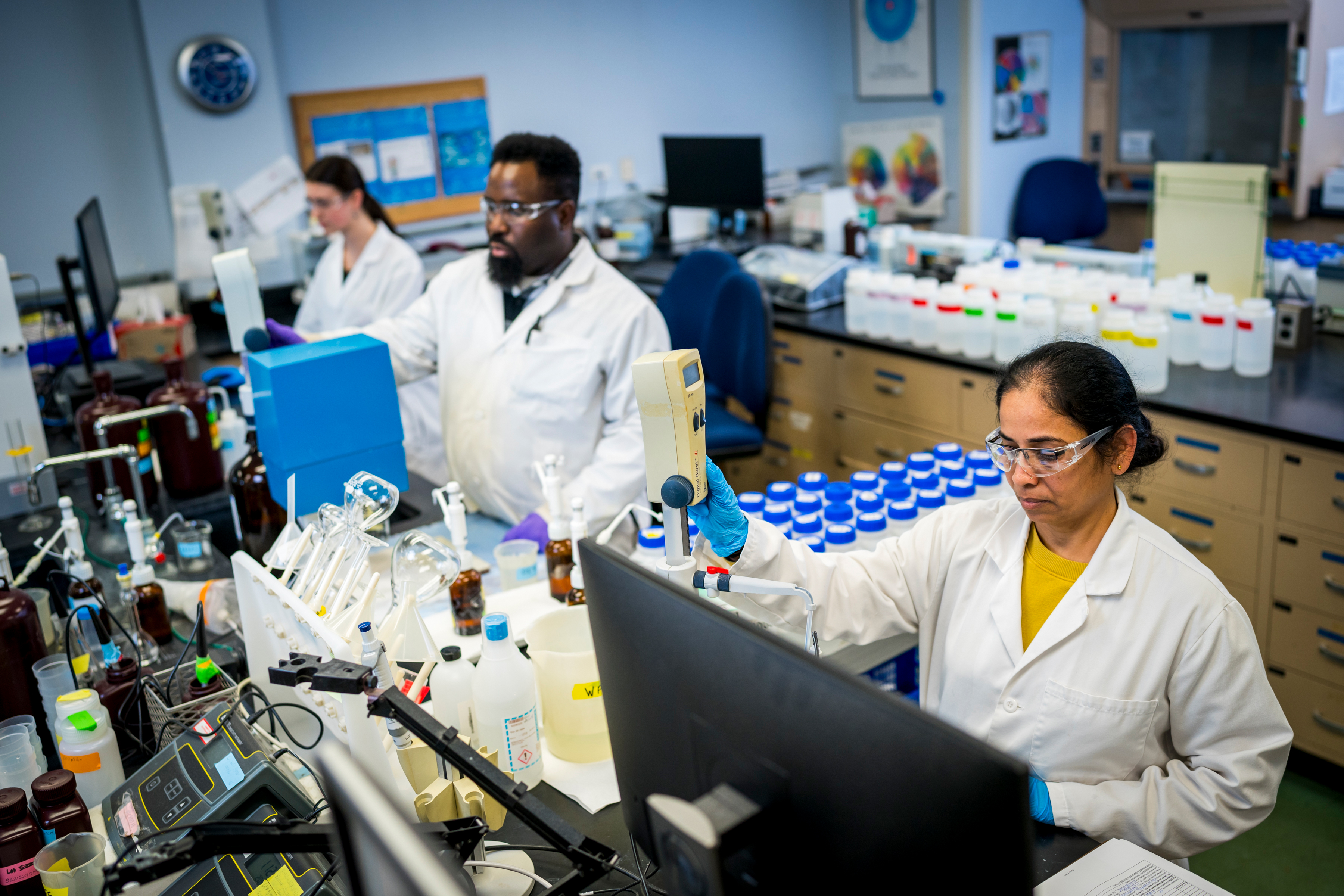 Three PWD scientists in our lab, all wearing white lab coats, safety glasses, and disposable gloves, analyze water samples. Nearest the camera, a woman with light brown skin and dark hair in a low bun operates machinery.