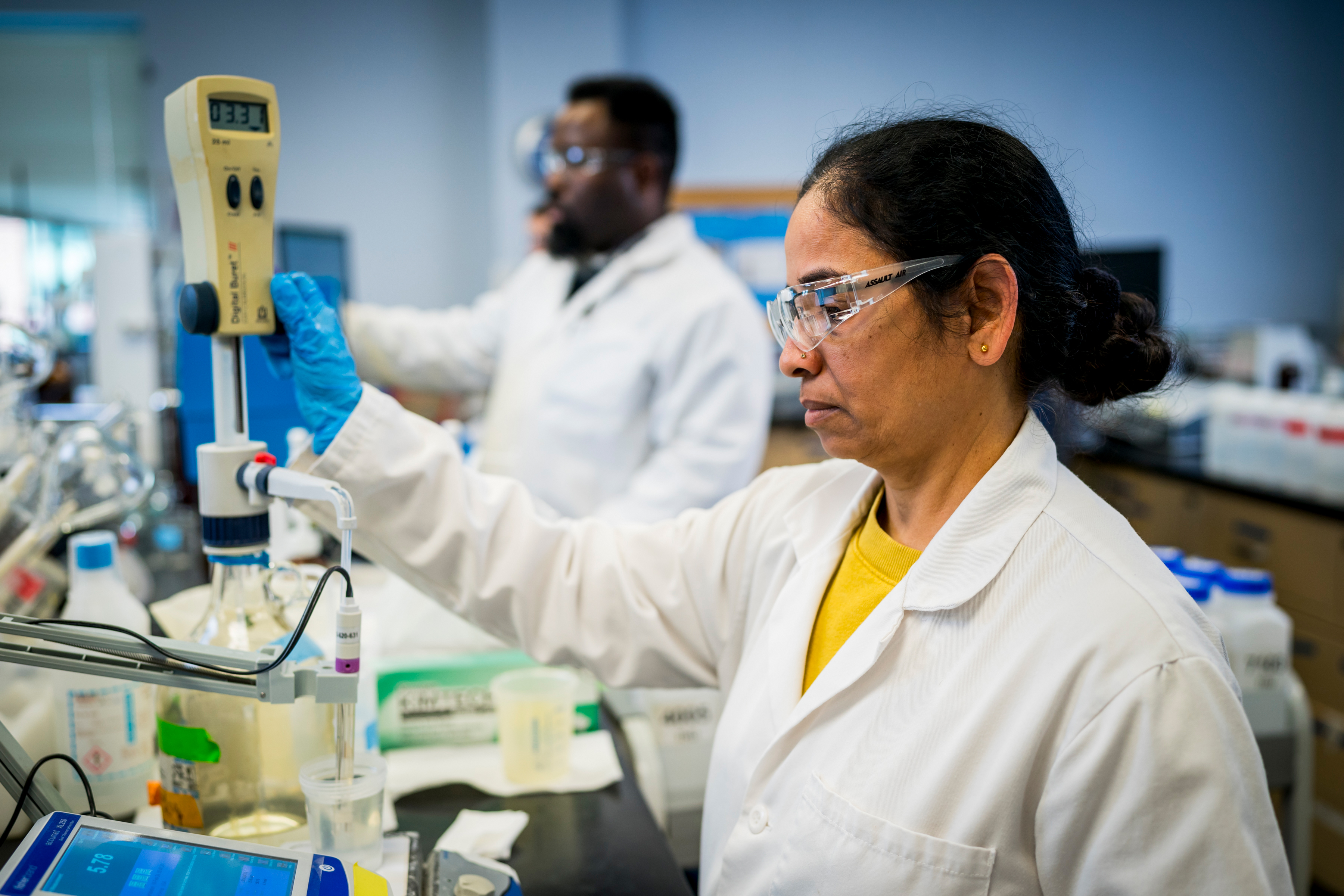 Two PWD scientists in our lab, wearing white lab coats, safety glasses, and disposable gloves, analyze water samples. Nearest the camera, a woman with light brown skin and dark hair in a low bun operates machinery.