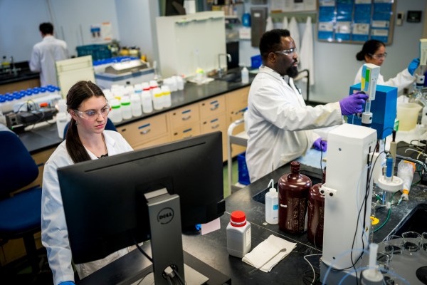 PWD scientists in our lab, all wearing white lab coats, safety glasses, and disposable gloves, analyze water samples. Nearest the camera, a woman with light skin and a long brown ponytail draped over her shoulder stands behind a computer monitor. Next to her, a man with dark skin, short curly black hair, and a goatee and a woman with light brown skin and dark hair in a low bun operate machinery. Another person with short dark hair and light skin is seen in the background facing away from the camera.