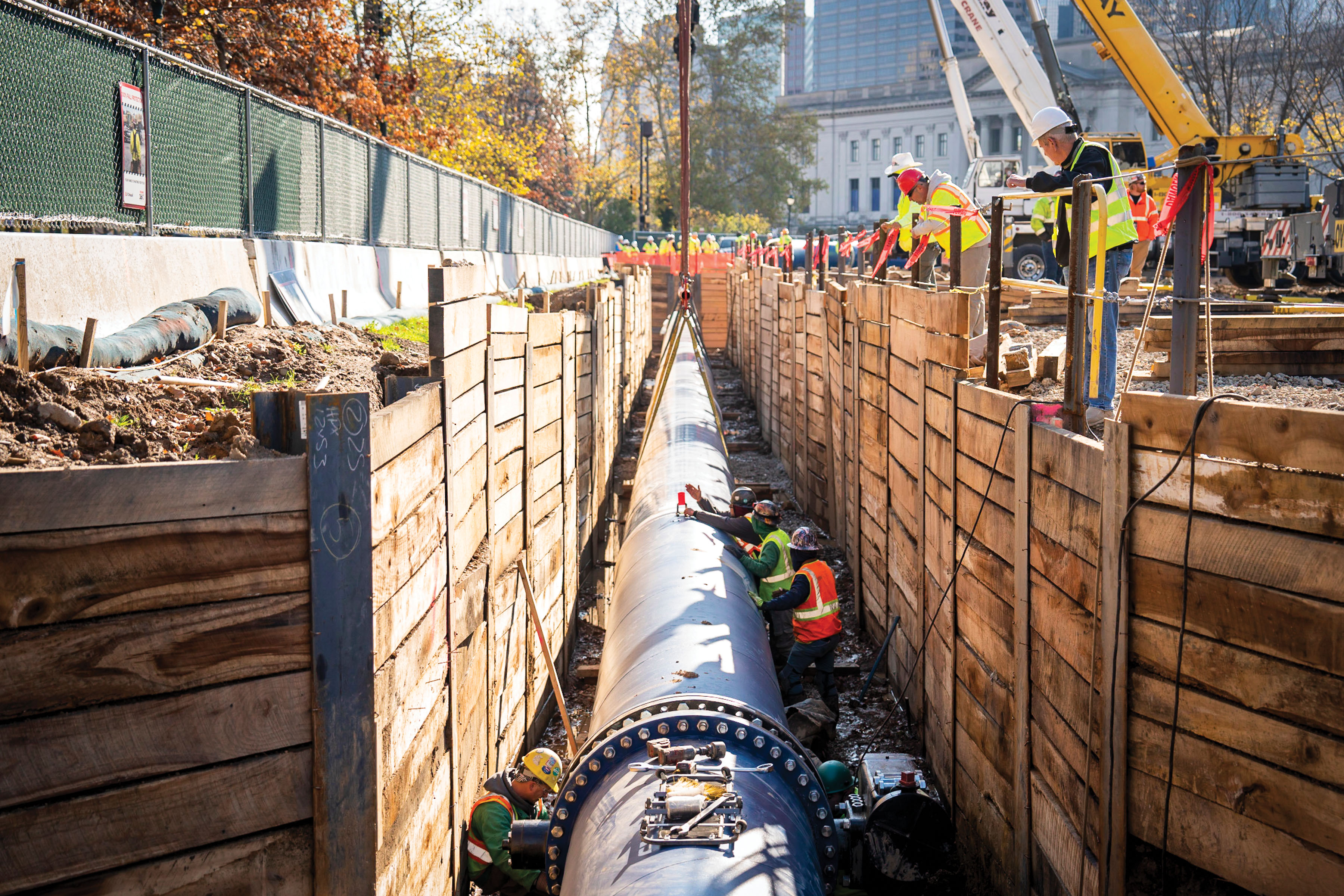 Workers secure a new section of water main that has just been lowered by crane into a large trench, stabilized with wood planks lining the sides.