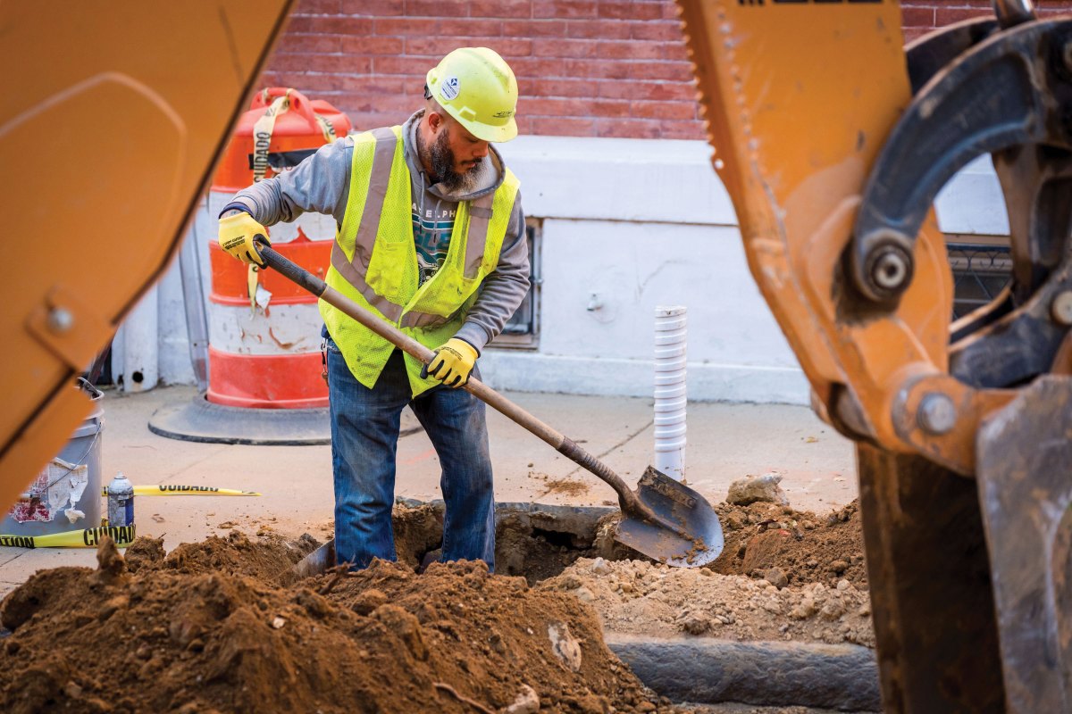 A worker wearing gloves, a hard hat, and reflective safety vest is seen from the knees up, standing in a hole in the sidewalk in front of a brick building, with a shovel poised to keep digging.