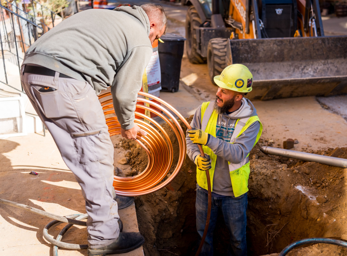 photo of two workers replacing a residential service line - one holds a coil of copper pipe as another, in neon yellow hard hat and vest, stands in a waist-deep hole in the sidewalk, straightening a section of the pipe to feed into position and install.