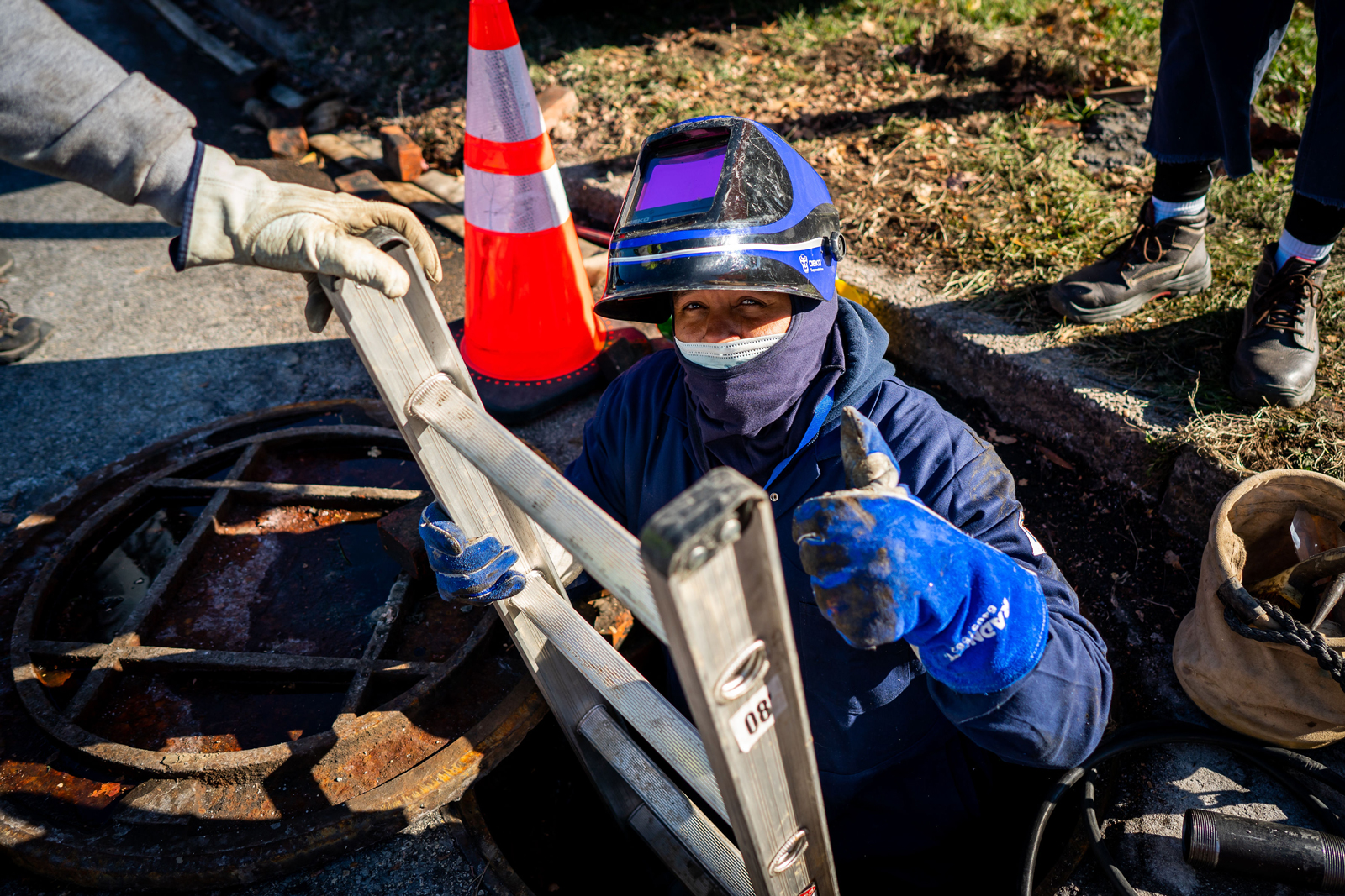 A PWD worker, geared up for both cold weather and safety in a winter coat, ski mask, work gloves, disposable face mask, and flipped-up welding mask, all in shades of blue, stands waist-high in a utility access hole, giving a thumbs-up while holding onto the ladder with the other. The hand of another worker is visible stabilizing the ladder, with the hole cover, an orange cone, various tools, and the feet of another worker seen in the background.