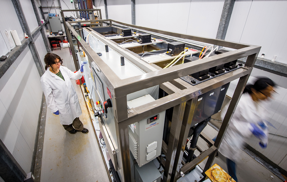 A photo taken inside the mini treatment plant container, from a high vantage point in one corner looking down the length towards the opposite corner, shows a large rack of equipment taking up nearly all of the space, leaving just enough space to walk around it on all sides. In the photo, two scientists, both with long, dark hair in ponytails, wearing white lab coats and purple gloves. One is on the left, doing something with the machinery, and the other is blurred, walking towards the right corner of the frame.