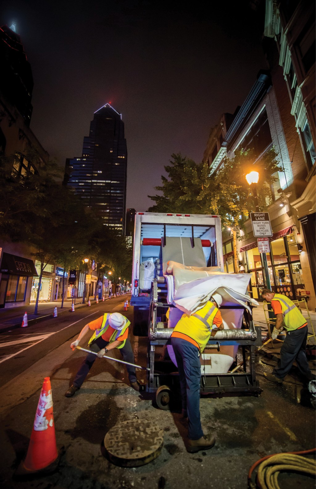 A PWD work crew wearing orange t-shirts, yellow safety vests, blue jeans, work boots, gloves, and white hardhats working on a sewer lining project in a closed-off Center City street at night.