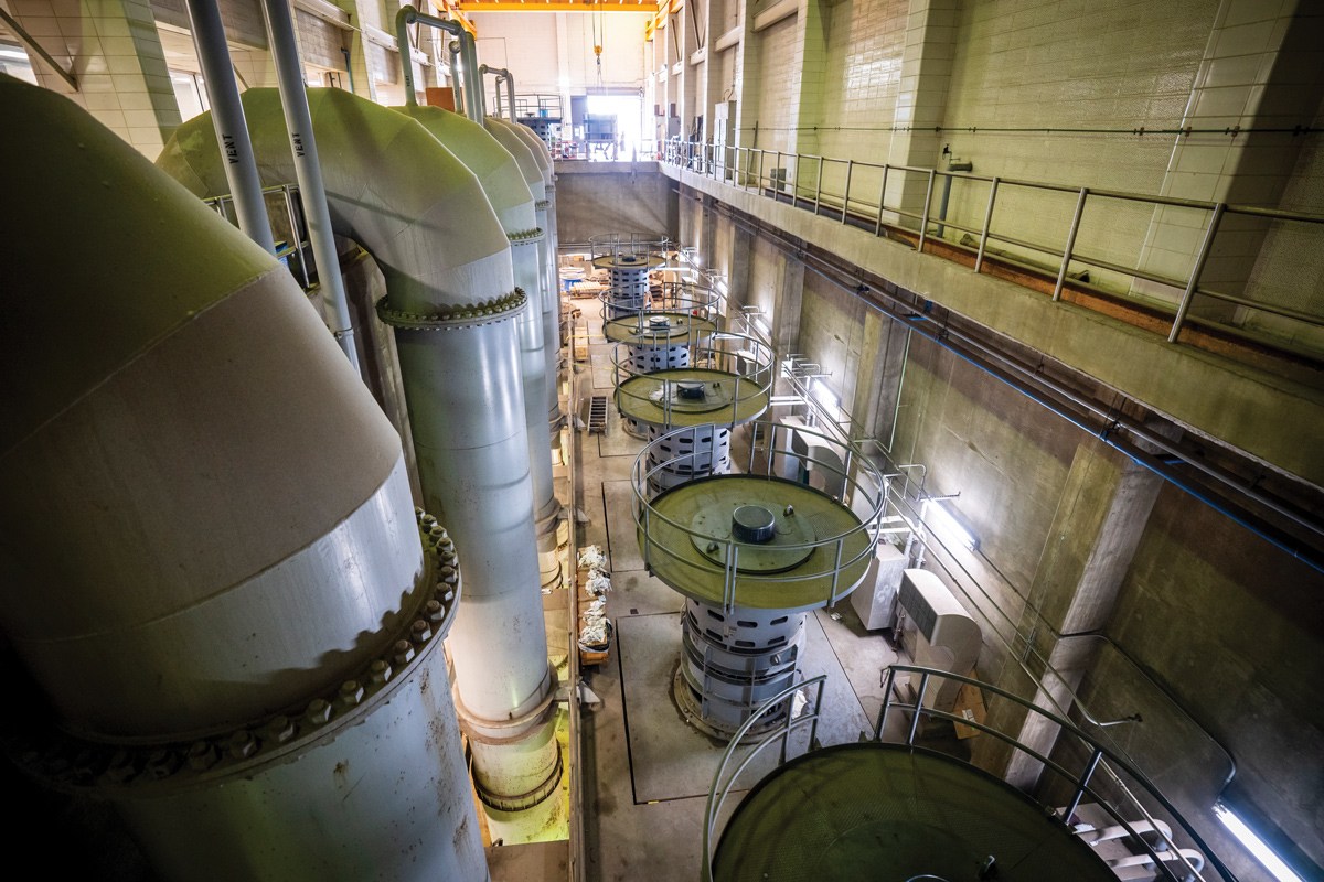 Interior view of a large room in our Northeast Water Pollution Control Plant. A row of huge arches of pipe fill the left side of the room, with, while round table shaped cylinders with hand rails around the top platforms fill the right side, and a walkway with a railing is visible above them on the right wall.