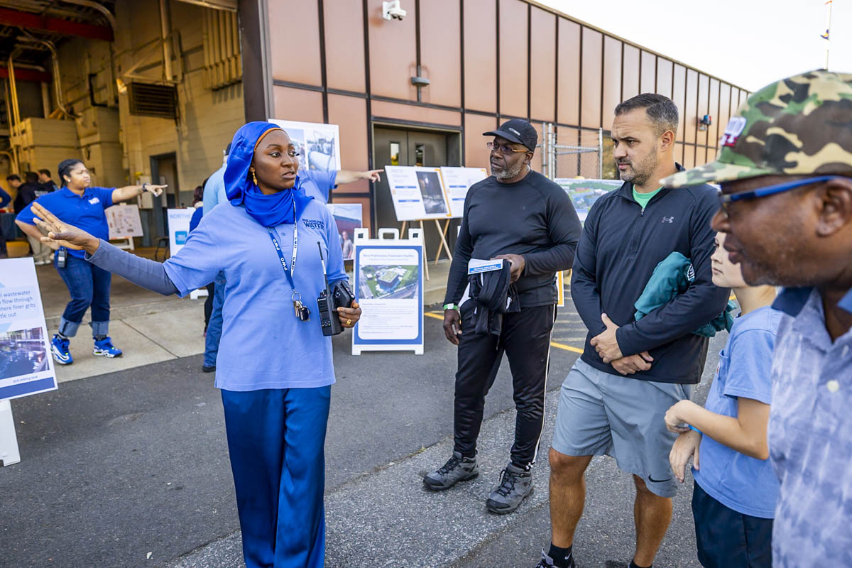 a PWD customer service representative wearing a bright blue hijab gestures while talking with several people in front of a boxy red-brown building with a large garage door open