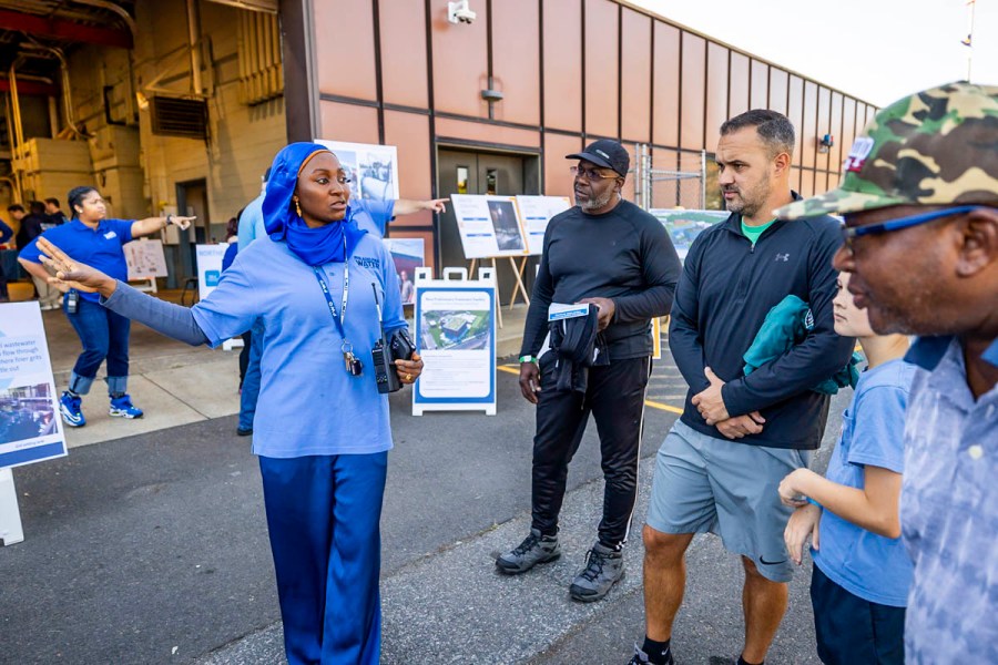 a PWD customer service representative wearing a bright blue hijab gestures while talking with several people in front of a boxy red-brown building with a large garage door open