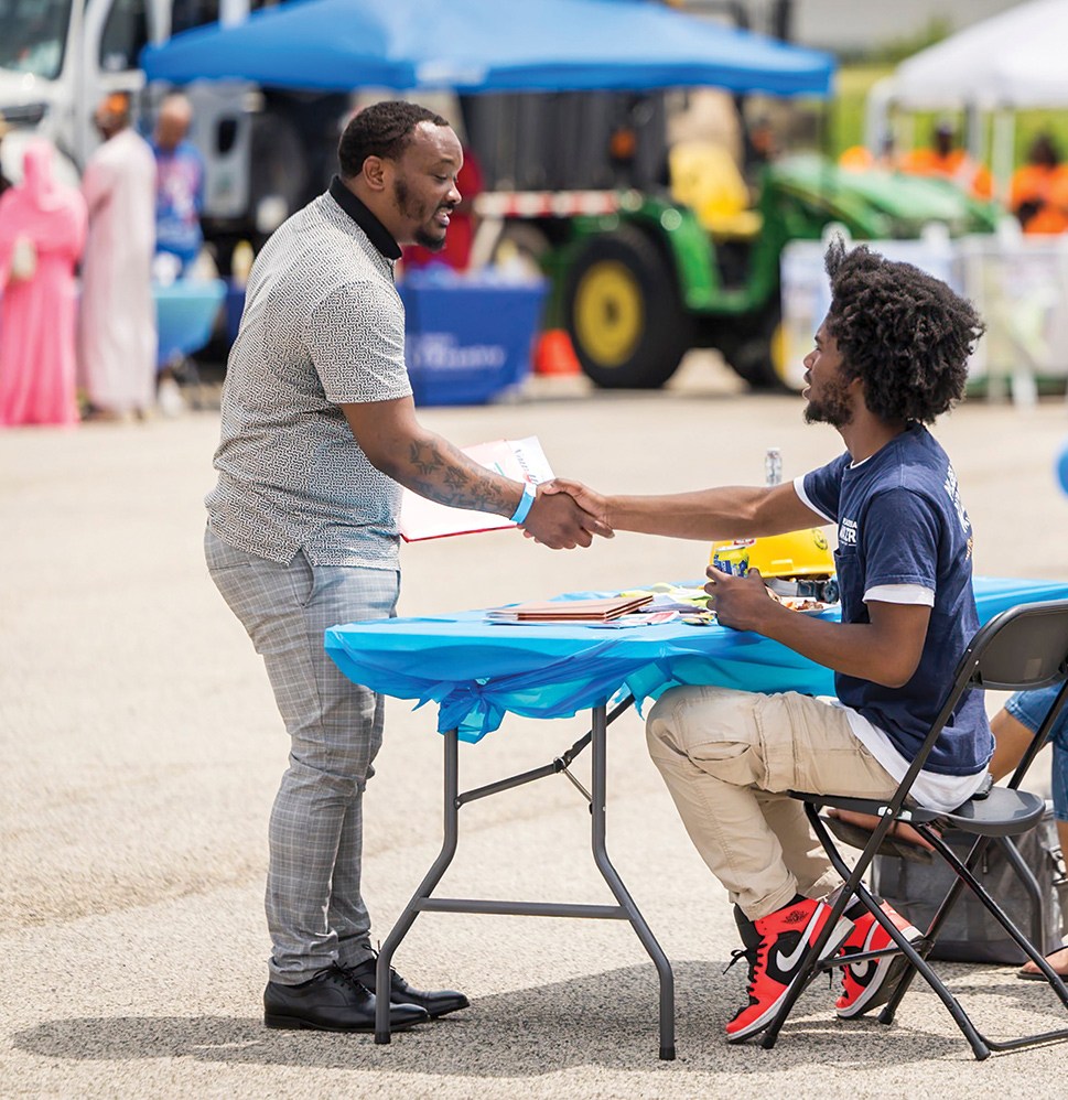 A guest arriving at an open house shakes hands with a PWD employee seated at a table. In the background a blue canopy and table with blue table cloth, a green tractor, and more guests are seen out of focus.