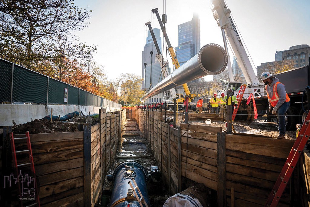 Multiple cranes lower a long section of water main into a trench with the sides stabilized by wooden planks. Workers in safety vests and hard hats are visible to the right of the trench, and skyscrapers are seen in the background.