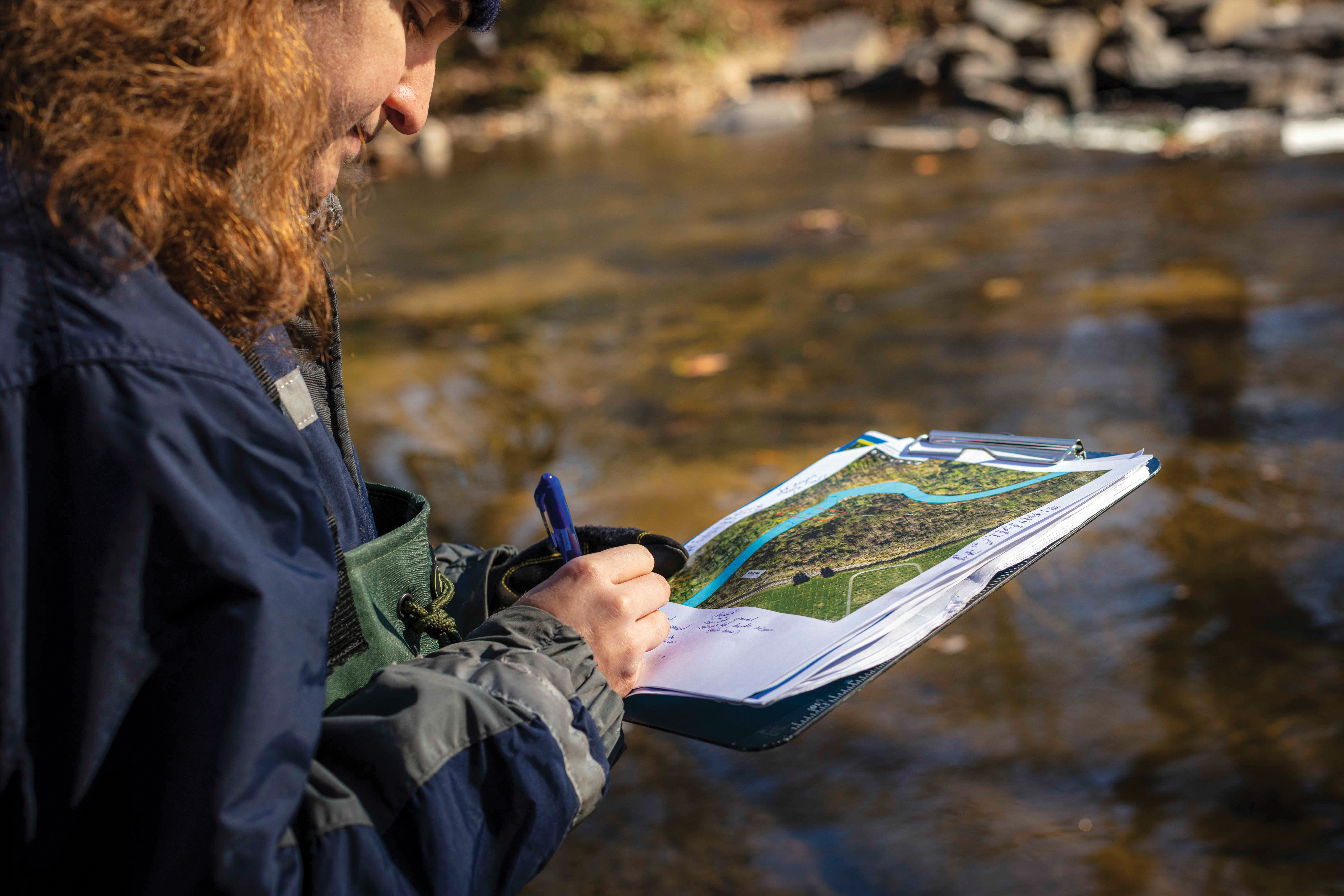 Looking over the shoulder of a PWD employee performing a stream assessment, writing notes on the bottom of a page on a clipboard featuring a map of the stream, with the actual stream blurry in the background.