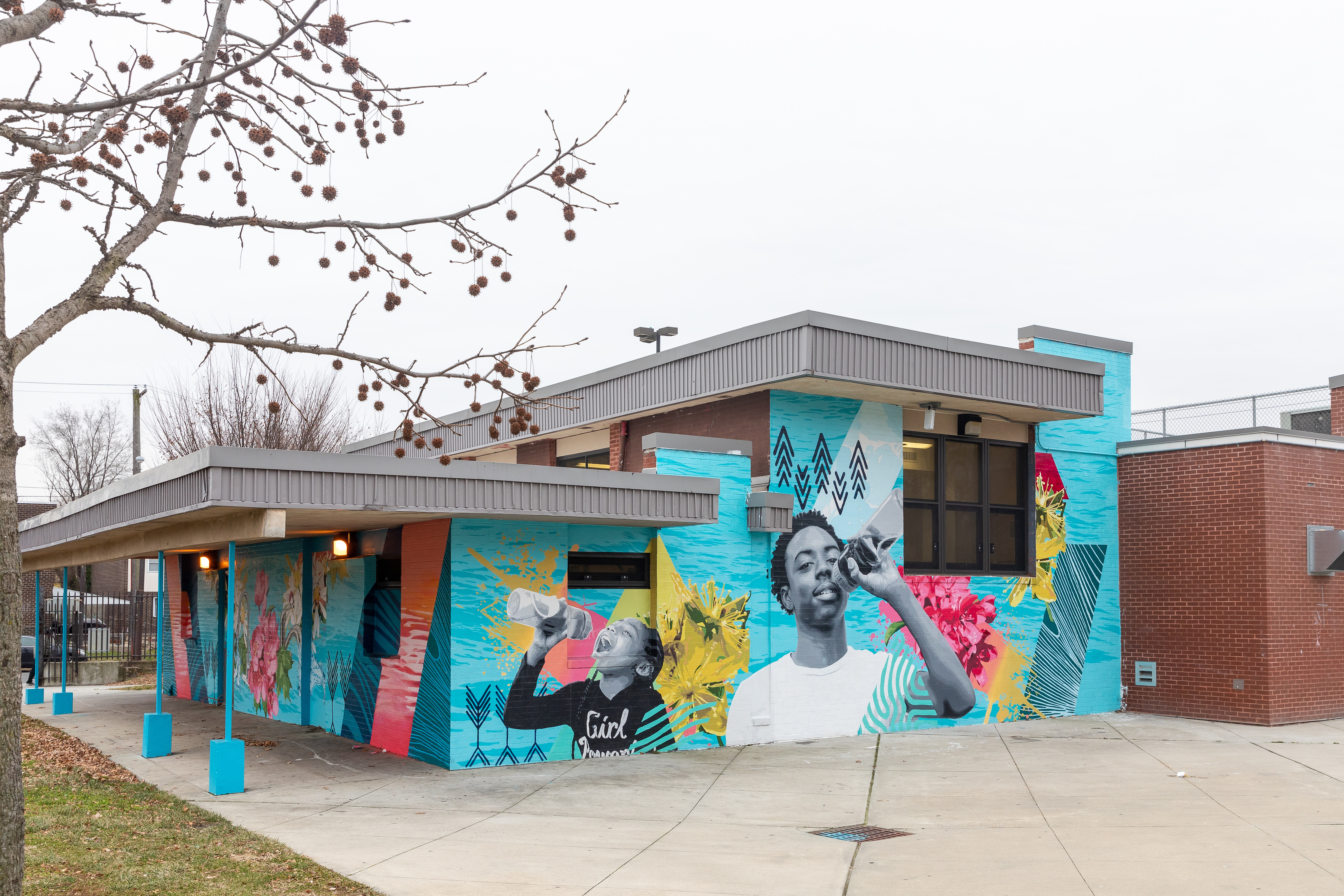 Two sides of a blocky brick building with multiple levels of flat roof are brightened by a bold and cheerful mural celebrating Philly's tap water - a background of bright turquoise water ripples overlaid with pink and yellow flowers and splashes and black and turquoise geometric patterns, and two grayscale portraits of a cool young black man in a white t-shirt and an excited and cheerful young black girl with braids, both sipping from reusable water bottles.