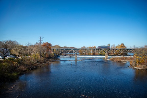 Looking upstream on the Schuylkill River between Bridgeport and Norristown, PA, as a single car on the Norristown High Speed Line crosses the train bridge. A couple people are seen standing in the water near the shore in front of the bridge.