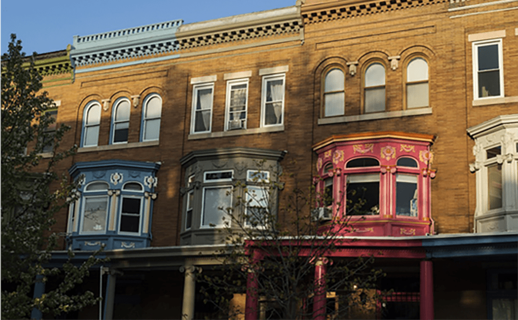 the fronts of a group of brick three-story rowhomes in Philadelphia, each with a front porch, a bay window on the second floor, and three narrow windows on the top floor, each with their trim painted a different color (blue, green, red, and cream)