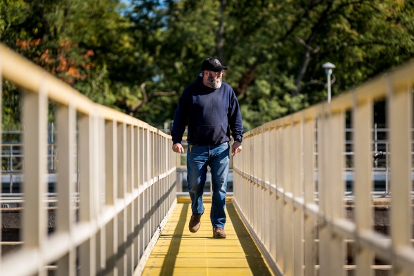 Treatment plant employee wearing a blue hoodie, jeans, work boots, and baseball cap, crosses the walkway over an outdoor settling basin, surveying the water below.