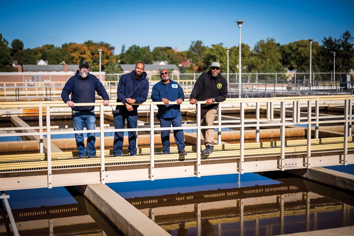 Four employees of our Queen Lane Drinking Water Treatment Plant, a mix of ages and races yet all sporting fine beards, pose leaning against a railing on a walkway over  sedimentation tanks.