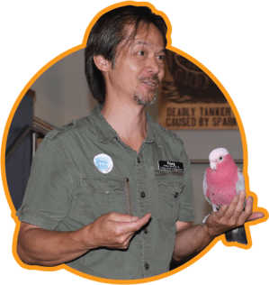 A man in a khaki shirt gestures with one hand while holding a bird with a white head and pink chest in the other.