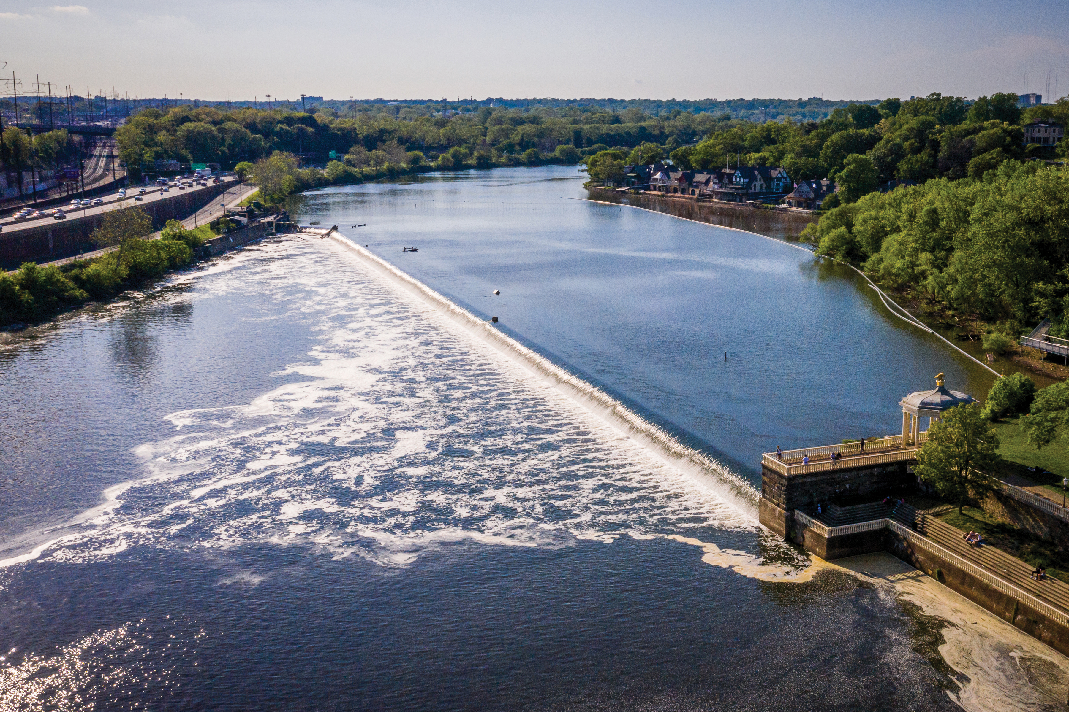 Looking upstream over the Schuylkill River, the Fairmount Dam stretches diagonally from the upper left to the lower right, creating a shallow waterfall. Boathouse Row is visible in the upper right and highway and train tracks on the left, with the trees of Fairmount Park and beyond in the background.