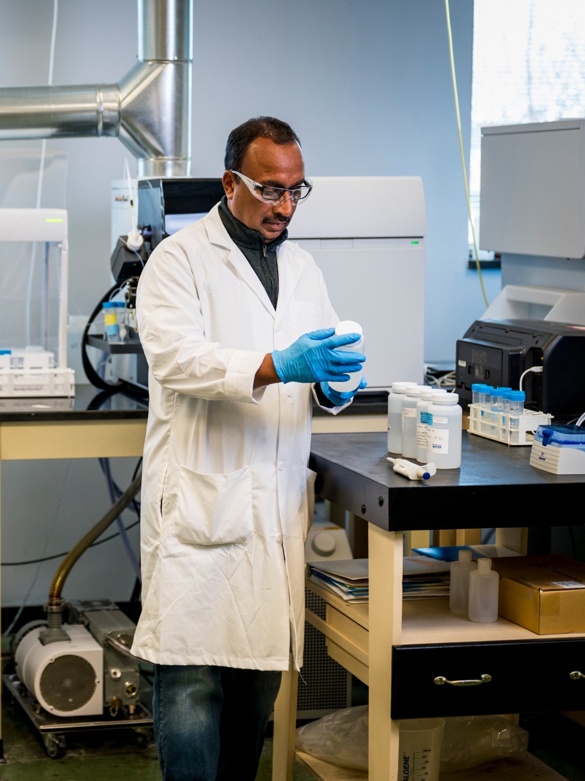 A scientist stands by a lab table, wearing safety glasses, a white lab coat, and blue disposable gloves, examining a water sample collection bottle, with several others lined up on the table and various machines visible in the background.