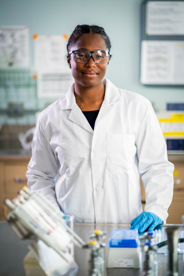 A scientist with dark textured hair in two braids, wearing safety glasses, a lab coat, and disposable gloves, looks across a lab table towards the camera with a small smile.
