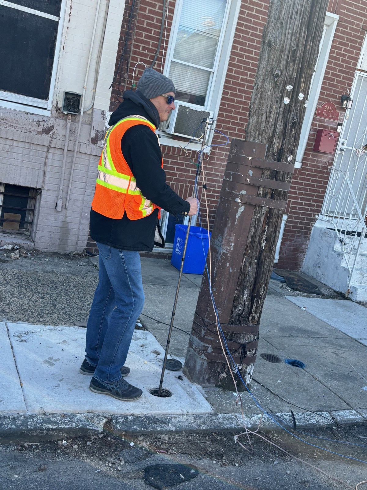 A contractor in a safety vest stands on the sidewalk in front of some row homes, holding a rod with wires connected to the top in a hole, using acoustic vibrations to check the material of a buried service line.