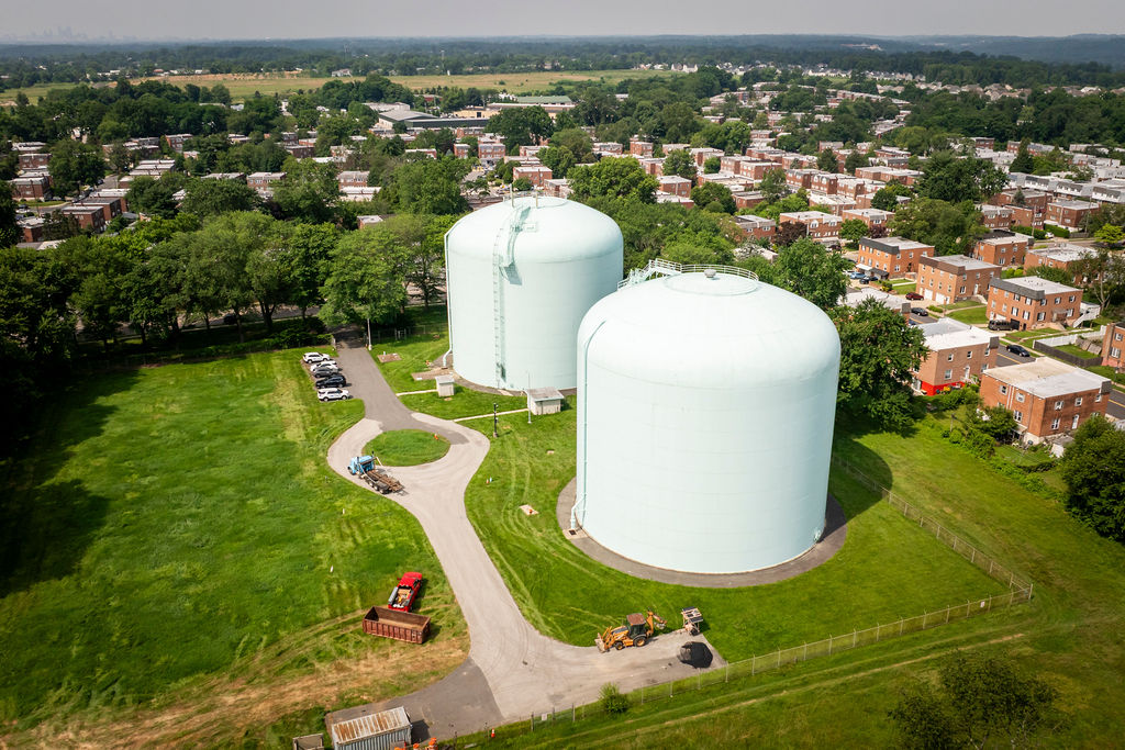 Two large light green water storage tanks next to a driveway with a cul-de-sac with houses and trees behind them.